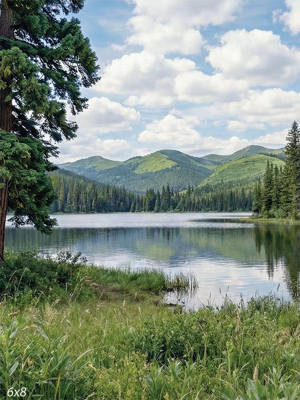 Printed photography backdrop showing a calm lake bordered by evergreen trees, grasses, and distant green mountains under a bright cloud-filled sky. The studio background has a quiet natural landscape with reflective water and layered forest detail.