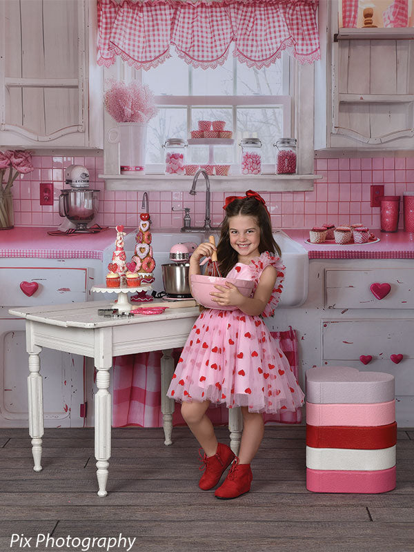 Young girl holds pink mixing bowl in a charming Valentine kitchen setup featuring red accents and heart-themed props.