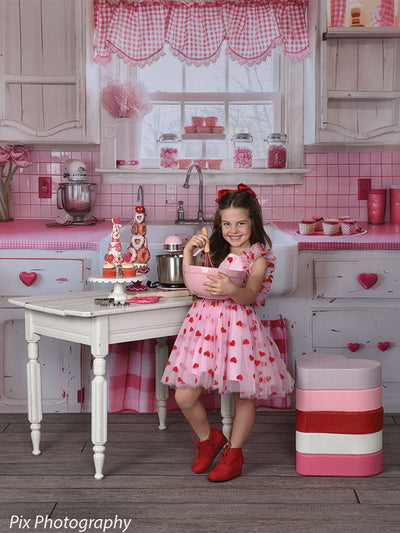 Young girl holds pink mixing bowl in a charming Valentine kitchen setup featuring red accents and heart-themed props.