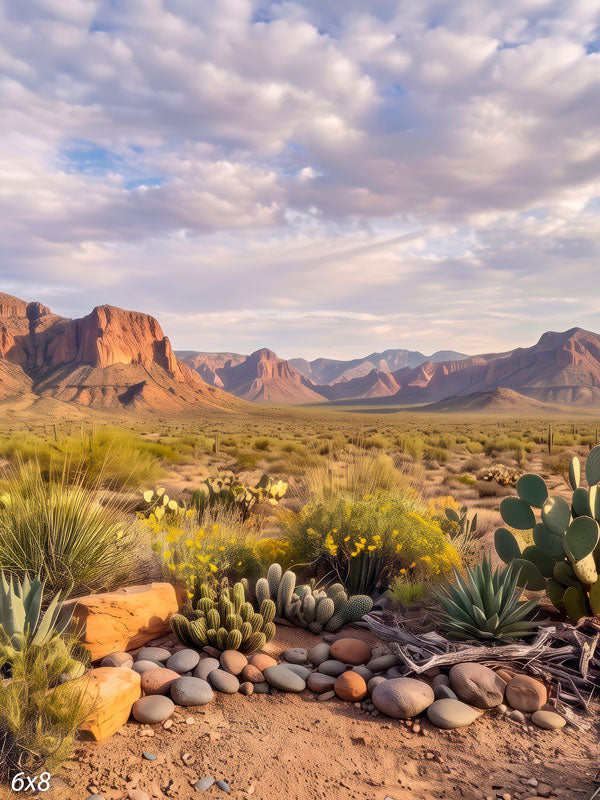 Desert photography backdrop with red rock mesas beyond a foreground of prickly pear, agave, golden wildflowers, and smooth stones under a soft cloudy sky.