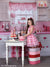 Girl stirs pink bowl in a playful heart-filled kitchen backdrop with pink gingham curtains and countertop treats.