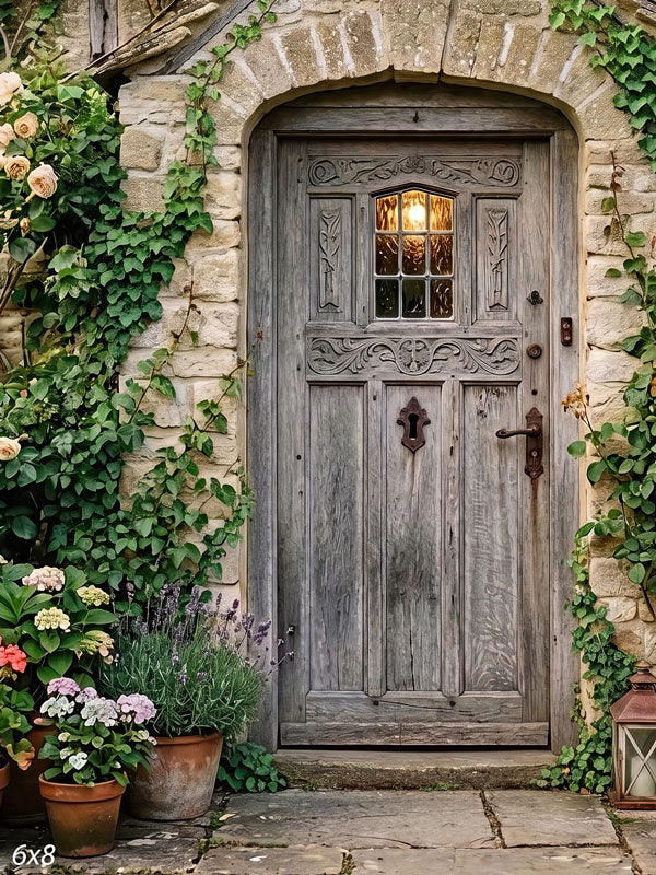 Arched weathered wood cottage door in a honey-colored stone wall with climbing ivy, cream roses and potted hydrangeas, a garden photography backdrop.