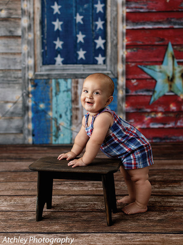 Baby boy in plaid romper standing and smiling beside a rustic dark wood photography prop stool, staged on a patriotic backdrop with vintage Americana styling.