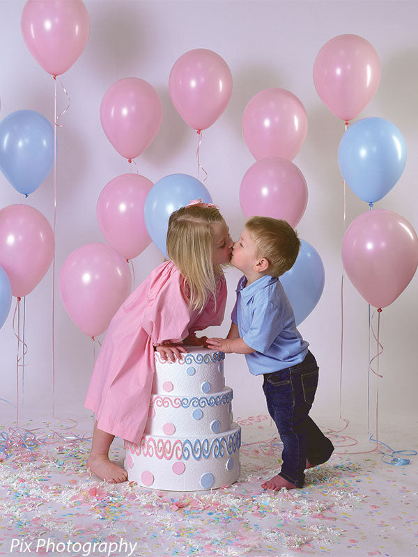 Light gray backdrop enhances colorful cake and balloons as kids share a kiss in a birthday photo session.