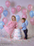 Young girl and boy pose in birthday scene with pastel balloons and cake, all set against seamless gray backdrop.