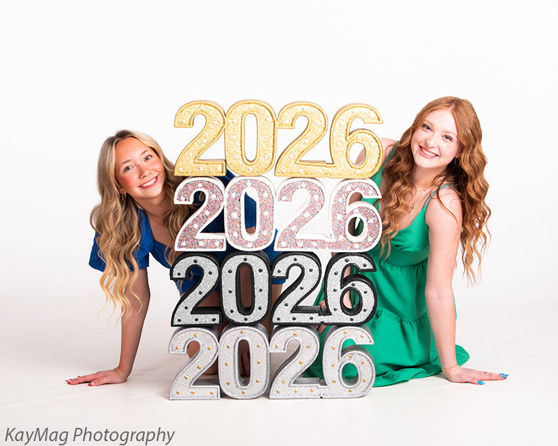 Seniors pose low beside stacked glitter “2026” prop, showing off creative graduation setups with fun, youthful energy.