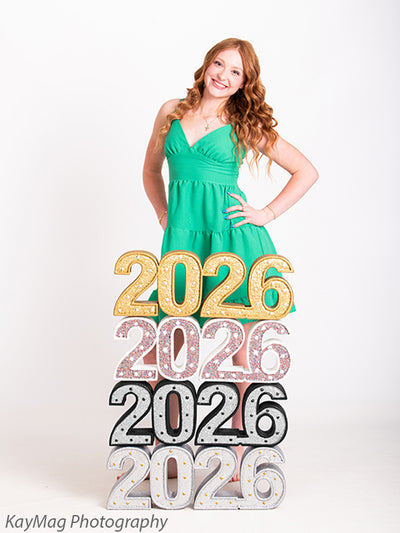 Red-haired senior girl in green dress smiles behind vibrant glitter “2026” prop, styled for graduation-themed studio portraits.