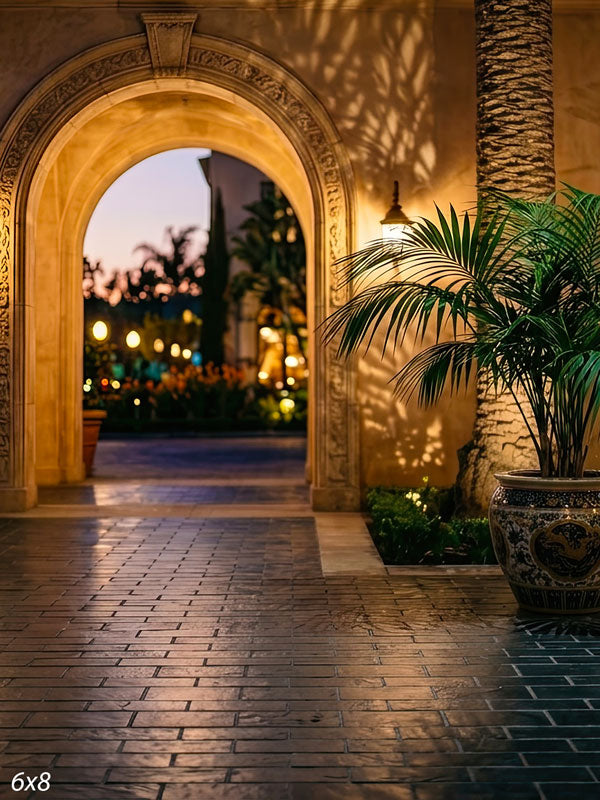 Warm stucco archway opening onto a twilight garden courtyard with a blue-and-white glazed urn, palm, and bokeh lights, a scenic photography backdrop.