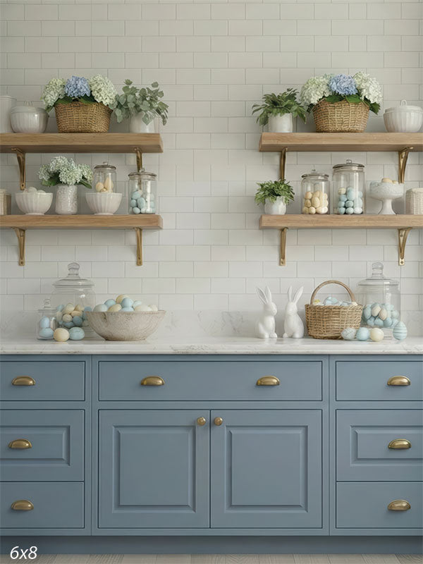 A photography backdrop depicting a symmetrical kitchen scene with tall blue cabinets and white subway tiles. Open shelves display hydrangeas and pastel eggs above a marble countertop.