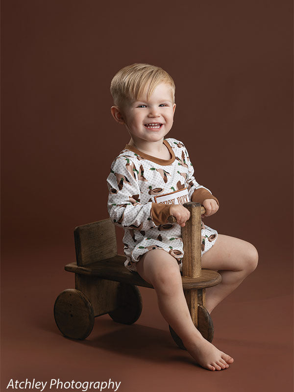 Joyful toddler posing on scooter prop in studio with brown backdrop, adding charm to milestone or themed portraits