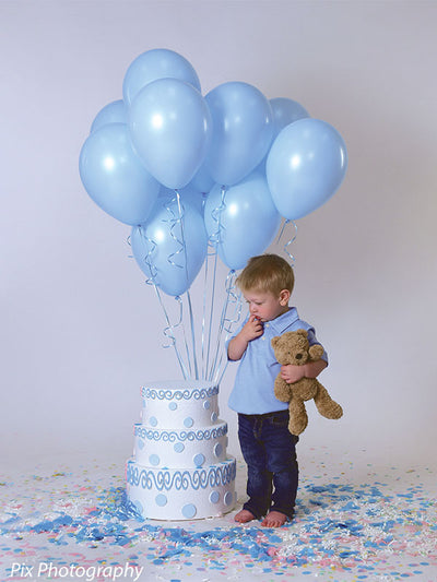 Boy with teddy bear stands against smooth light gray backdrop with pastel blue balloons and birthday cake prop.