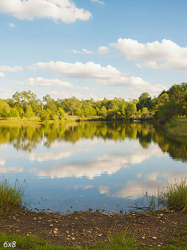 Calm lake mirroring a blue sky with puffy clouds, framed by a green tree line and grassy banks, an outdoor photography backdrop.