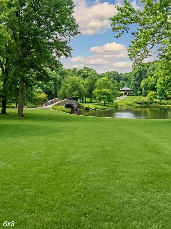 Groomed park lawn sloping to a pond with an arched stone bridge and a gazebo framed by mature trees, a scenic photography backdrop.