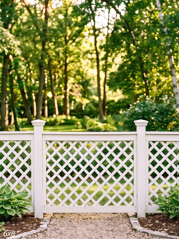 White lattice garden fence with square-capped posts standing in front of a sunlit green tree canopy with hostas along a gravel path, a photography backdrop.