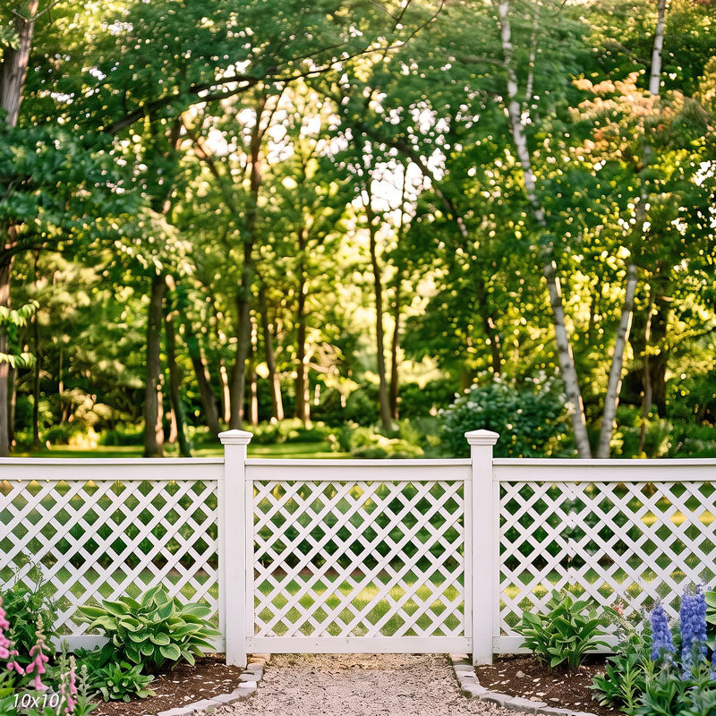 Wider view of the white lattice fence showing more of the summer tree canopy and the flower beds along the gravel path, a garden photography backdrop.