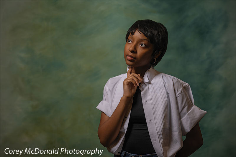 Green abstract hand-painted photography backdrop shown in a wide studio senior portrait with a young woman resting a hand at her chin, looking upward OM-2644