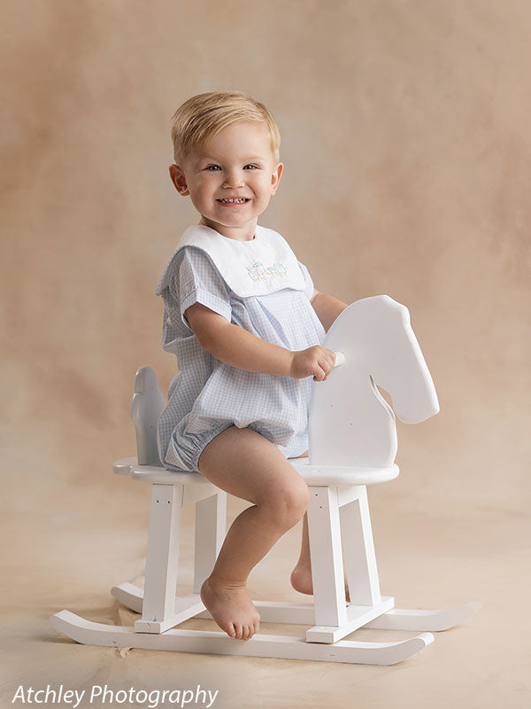 Happy toddler posing with handcrafted white horse photography prop, evoking classic childhood joy and studio warmth