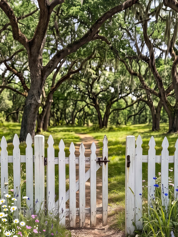 Open white picket gate in a low fence with a dirt path leading into a moss-draped live oak grove and wildflowers along the base, a photography backdrop.