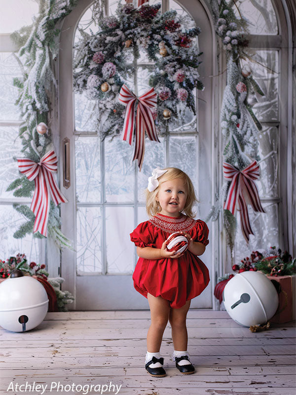 A charming winter-themed photography backdrop showcasing a beautifully decorated arched door with snow-kissed garlands, red berries, and holiday ribbons.