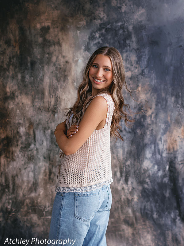 Young woman with long wavy brown hair turned slightly over her shoulder, wearing a cream knit sleeveless top and light blue jeans, smiling in front of an abstract brown earth texture photo backdrop with layered gray and warm brown tones.