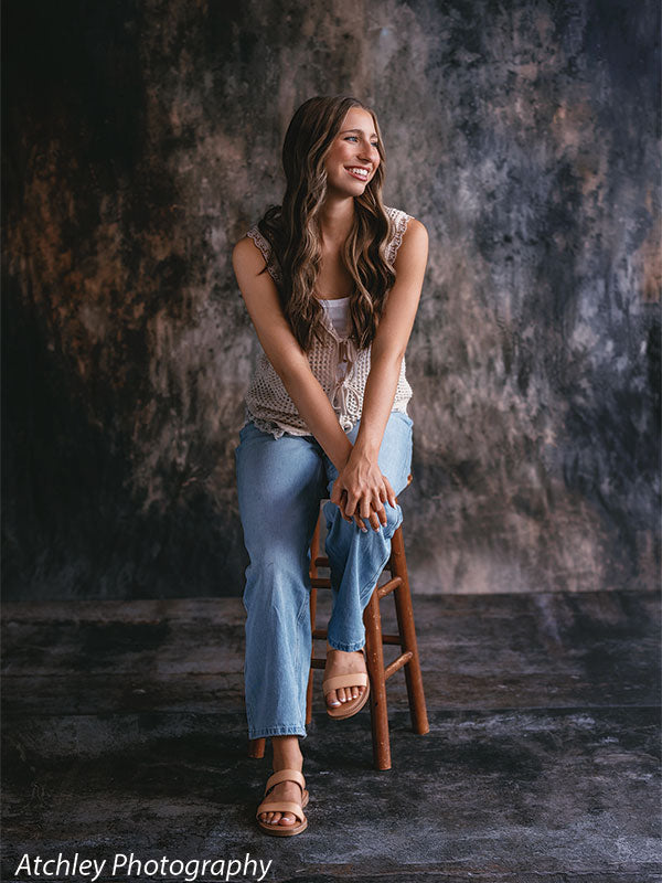 Young woman with long wavy brown hair sitting on a wooden stool wearing a cream knit sleeveless top, light blue jeans, and tan sandals, posed in front of an abstract brown earthscape printed backdrop with blended gray and warm brown tones and a coordinating concrete floor.