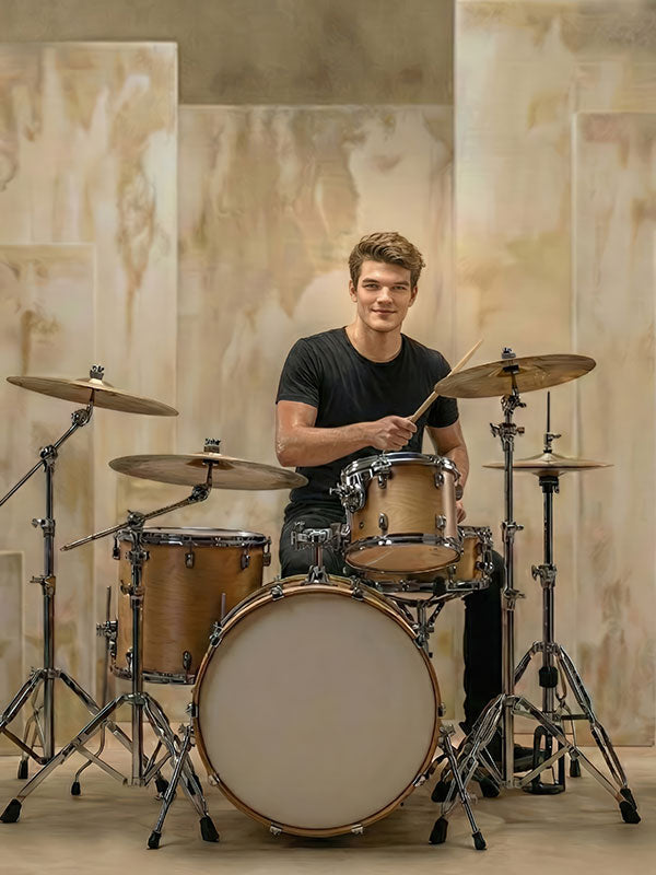 Man wearing a black shirt sits at a wooden drum kit holding drumsticks, positioned in front of a beige abstract painted panel backdrop with layered vertical panels.