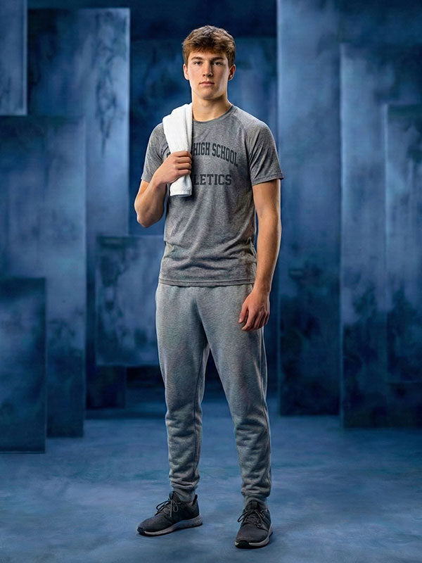 Young man wearing a gray athletic t-shirt and sweatpants holds a white towel over his shoulder while standing against a blue abstract painted panel backdrop with layered blocks. 