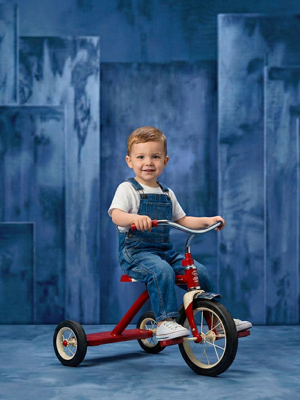 Smiling toddler boy wearing denim overalls and a white shirt sits on a red tricycle against a blue abstract painted panel backdrop with staggered rectangular panels.