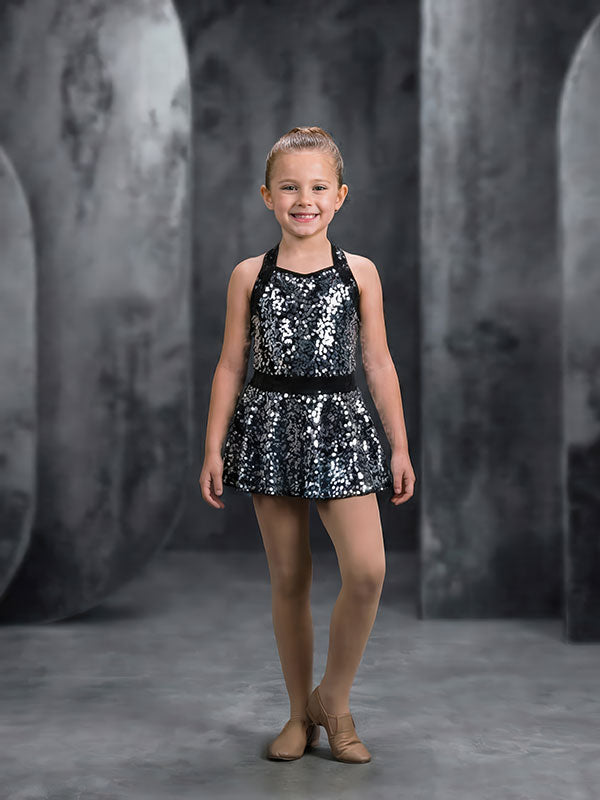Young girl wearing a black sequined dress and tan ballet shoes stands with one foot slightly forward against a gray abstract painted panel backdrop with curved and rectangular textures.