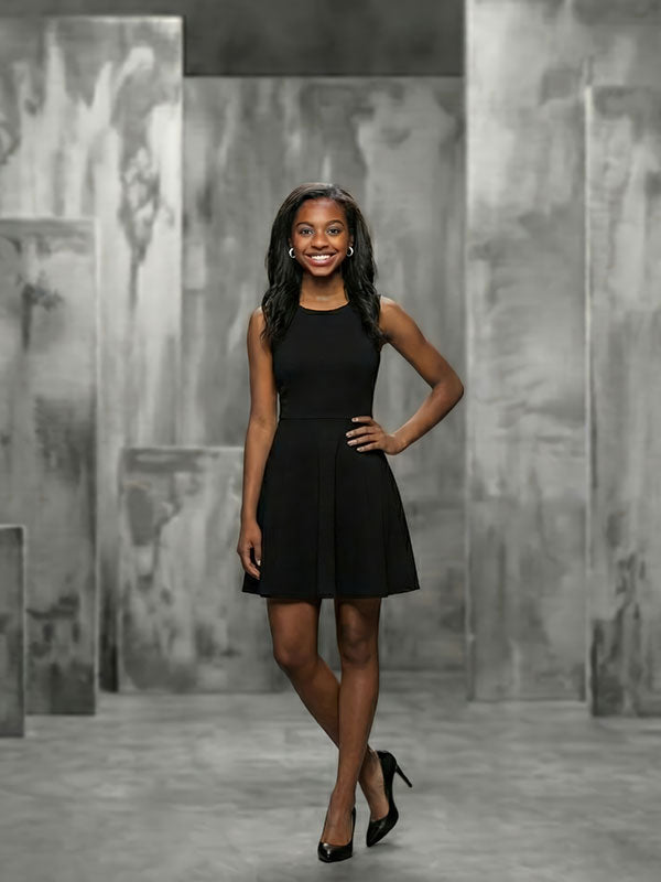 Woman wearing a black sleeveless dress and black heels stands with one hand on her hip, smiling in front of a gray abstract painted panel backdrop with layered vertical panels.