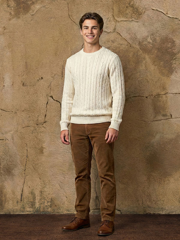 Young man wearing a cream cable knit sweater and brown pants stands smiling with arms relaxed at his sides in front of an abstract textured painted wall backdrop with warm neutral tones and visible cracks.