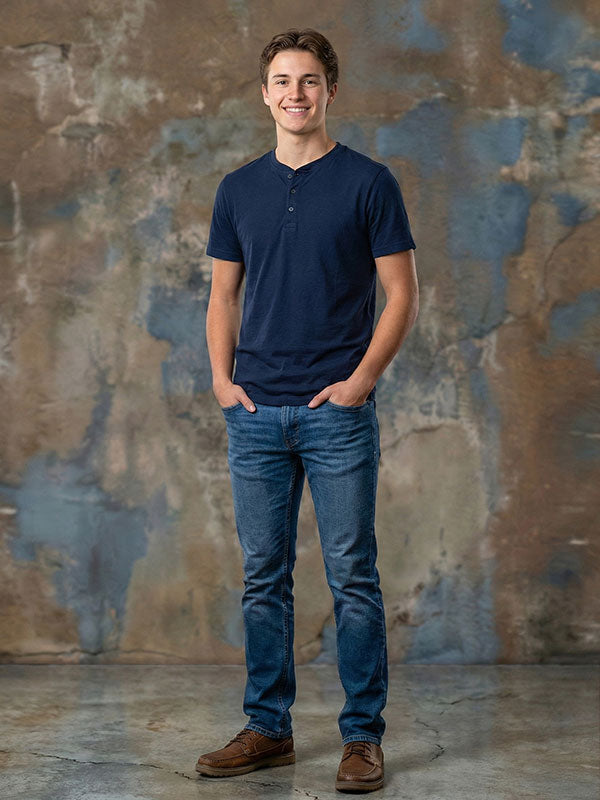 Young man wearing a navy short sleeve shirt and blue jeans stands casually with hands in pockets in front of an abstract textured painted wall backdrop with soft neutral and blue tones.