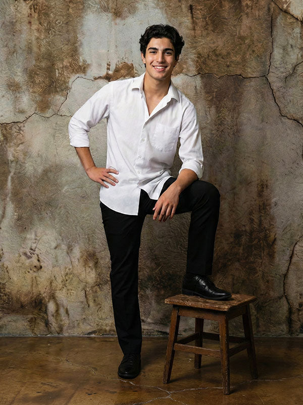 Young man wearing a white button-down shirt and black pants poses with one foot resting on a wooden stool, smiling in front of an abstract textured painted wall backdrop with warm brown and beige tones.