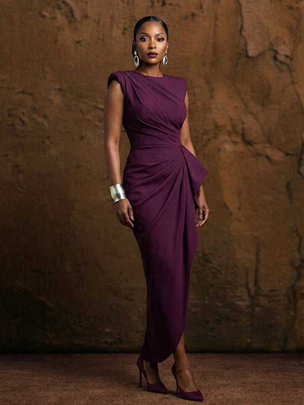 Woman wearing a structured deep purple dress with silver jewelry stands confidently with arms relaxed at her sides in front of an abstract textured painted wall backdrop with warm brown tones.