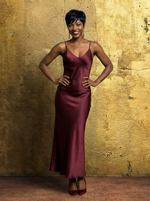 Woman wearing a burgundy satin dress stands smiling with hands on her hips in front of an abstract textured painted wall backdrop with golden yellow tones and subtle surface texture.
