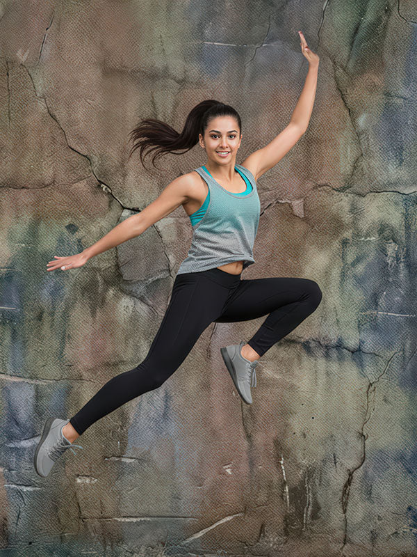 Athletic woman wearing a gray tank top and black leggings is captured mid-leap with arms extended against an abstract textured painted wall backdrop with layered neutral and blue tones.