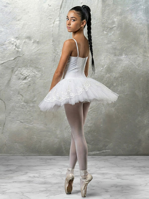Young ballet dancer wearing a white tutu and pointe shoes stands turned slightly away while looking over her shoulder, posed against an abstract textured painted wall backdrop in soft gray tones with a light marble-style floor.