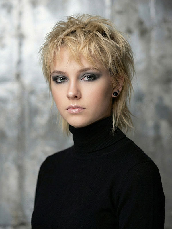 Woman with short blonde hair wearing a black turtleneck top faces forward with a neutral expression against an abstract textured painted wall backdrop with soft gray and muted neutral textures.