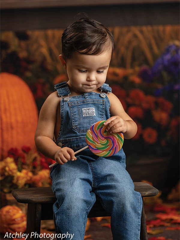 Smiling toddler in denim overalls sitting on a small rustic wooden bench with pumpkins and scattered fall leaves at his feet, in front of an autumn fairground carousel backdrop filled with flowers, gourds, and glowing carnival lights.