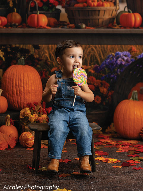 Happy toddler seated on a rustic wooden bench holding a rainbow lollipop, with pumpkins, mums, and fallen leaves around him, set against an autumn fairground harvest carousel backdrop with warm glowing lights.