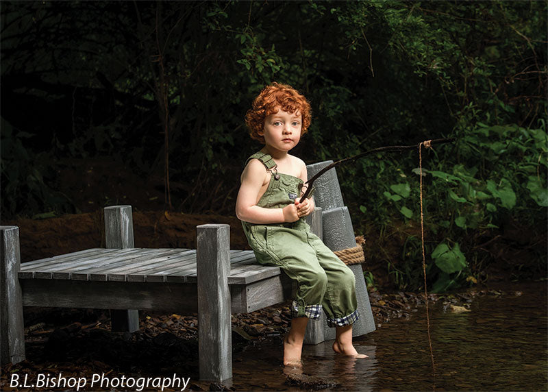 Beach piling photography prop shown with a red-haired toddler in green overalls seated on the gray wooden pier holding a twig fishing pole with bare feet in the creek SPL-3
