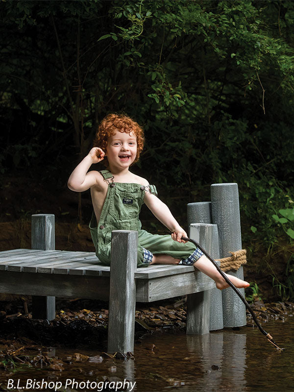 Beach piling photography prop shown with a red-haired toddler in green overalls grinning with a leg outstretched toward the gray piling posts and a fishing pole in hand SPL-3