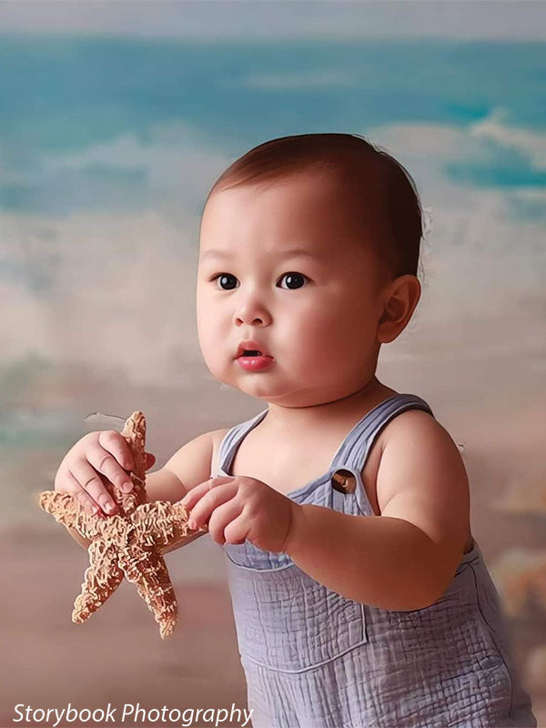 Infant portrait with coastal backdrop featuring sandy shore and waves, ideal for newborn and toddler beach-themed photography.