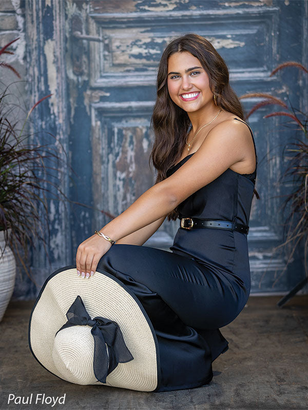 Black Ruin photography backdrop showing a weathered rustic door with peeling paint and an aged brick surround, shown with a senior girl in a black jumpsuit crouching while holding her wide-brim sun hat CP-8789
