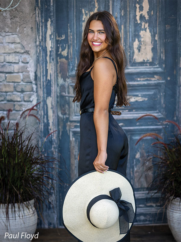 Black Ruin photography backdrop showing a weathered rustic door with peeling paint and an aged brick surround, shown with a senior girl in a black jumpsuit turned to the side holding her sun hat, laughing CP-8789
