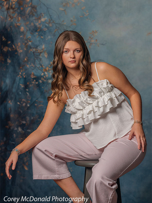 Teen girl with long wavy light brown hair wearing a white ruffled top and pink pants sits on a stool with one arm extended and a serious expression in front of a blue artistic backdrop with layered branch textures.