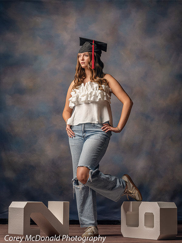 Teen girl with long wavy light brown hair wearing a white ruffled top, ripped jeans, and a graduation cap stands in a side pose with one leg raised on a number prop, looking to the side in front of a blue artistic backdrop with cloud texture effect.