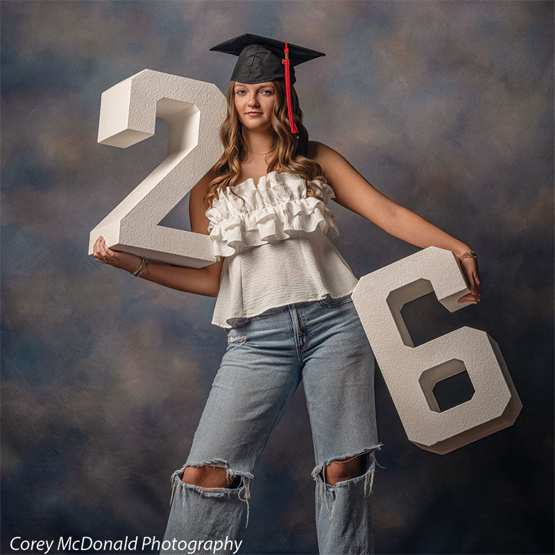 Teen girl with long wavy light brown hair wearing a white ruffled top, ripped jeans, and a graduation cap holds large white numbers two and six while looking directly forward with a neutral expression in front of a blue artistic backdrop with cloud texture effect.