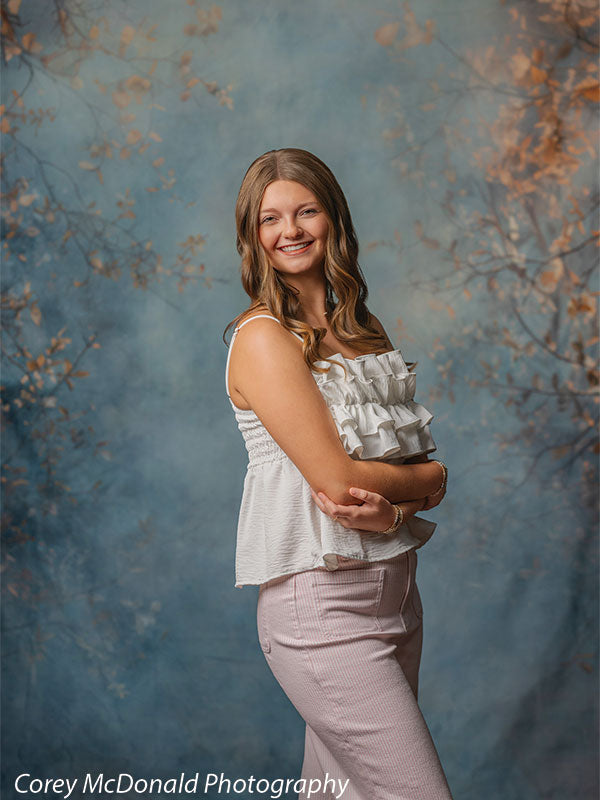 Teen girl with long wavy light brown hair wearing a white ruffled sleeveless top and light pink pants stands in a side pose with arms crossed, smiling in front of a blue painted backdrop with soft autumn leaf branches.