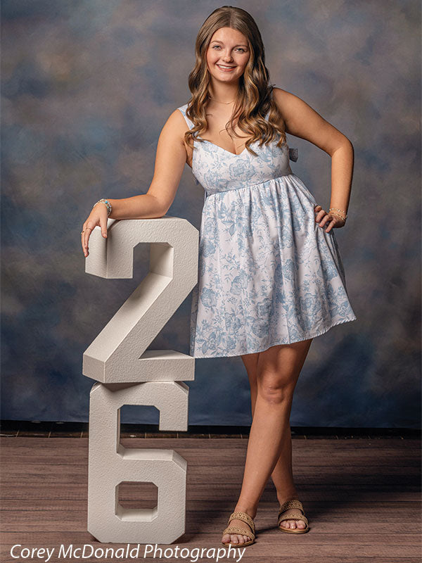 Teen girl with long wavy light brown hair wearing a light blue floral dress leans on a large white number two and smiles in front of a blue cloud texture photography backdrop.